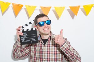 Smiling man wearing sunglasses holds a film clapperboard and gives a thumbs up in front of festive triangular banners.